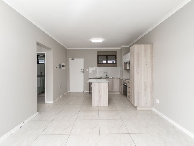 An open-plan kitchen area featuring light wood-finish cabinetry, a central island, tiled flooring, and integrated appliances, leading to an adjacent bathroom.