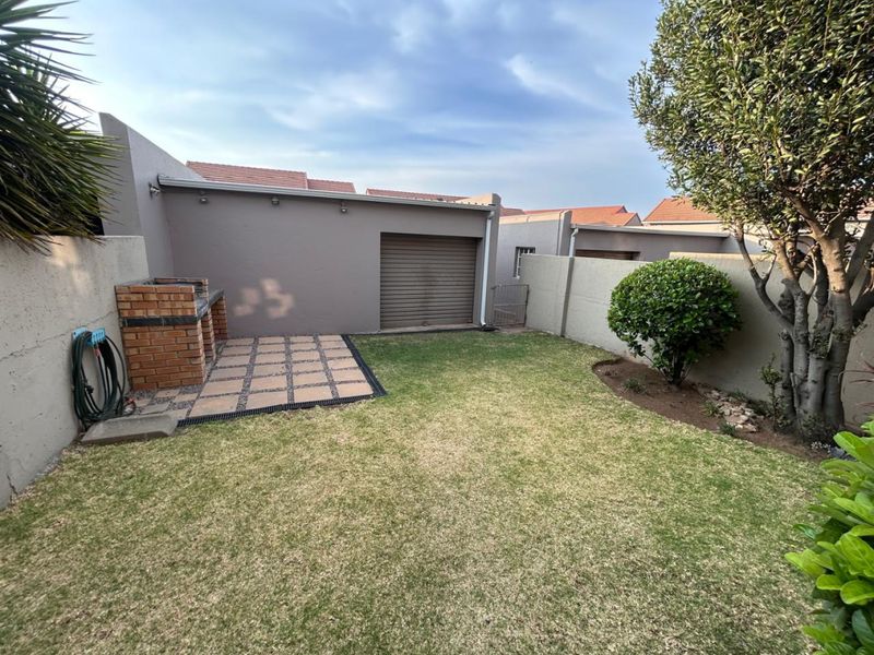 A private rear garden featuring a paved patio area with a built-in brick braai, a well-maintained lawn, and access to the garage.