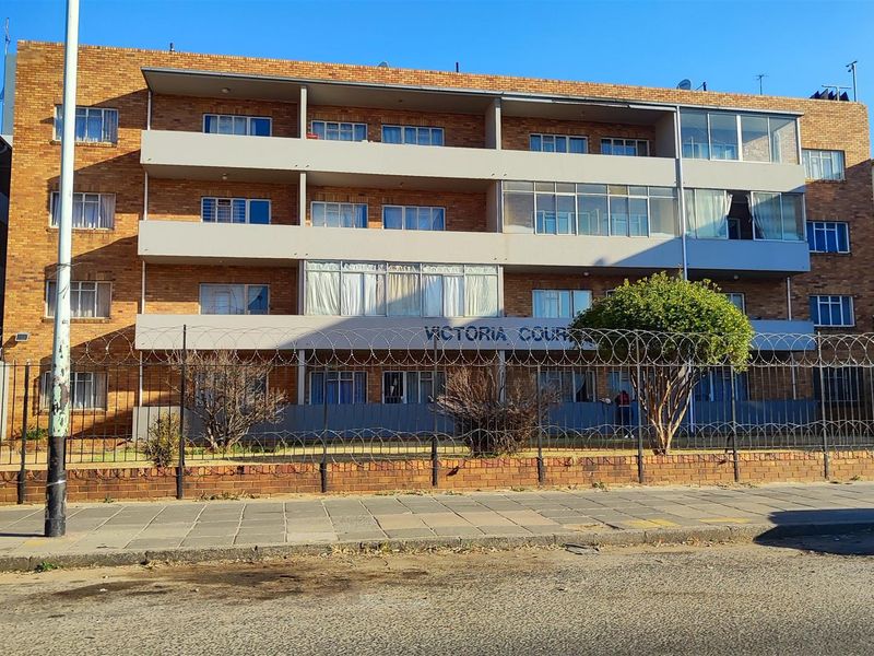 Street-facing view of Victoria Court, a multi-story apartment building featuring face brick construction, private balconies, and perimeter security fencing.