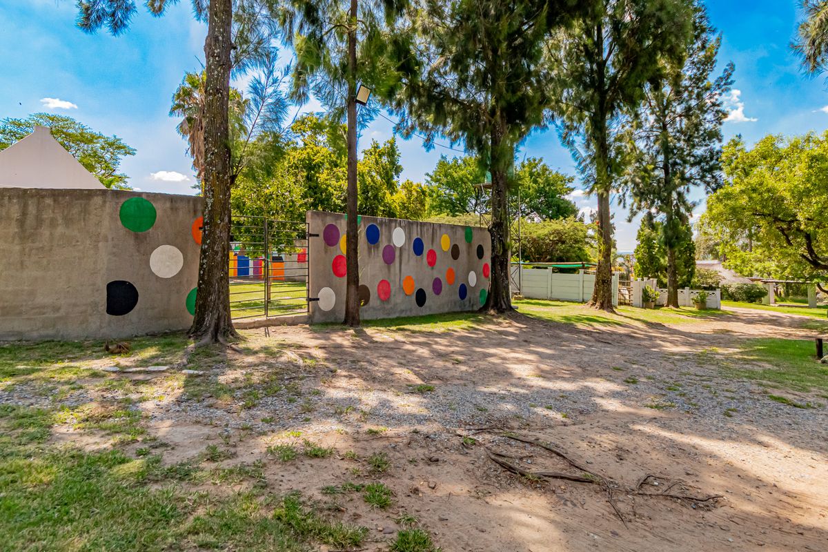 A view of the rear property grounds featuring a painted perimeter wall, metal gate, and mature trees on a gravel and grass surface.