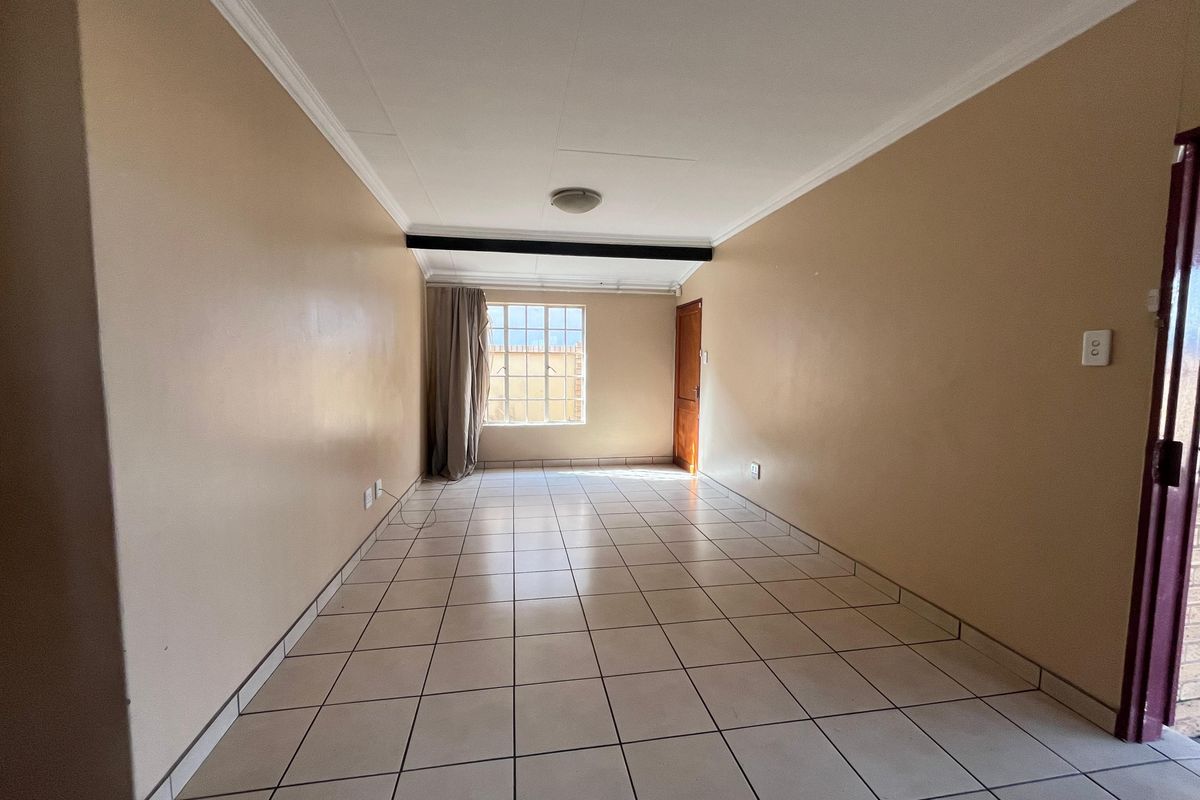 An empty, rectangular living area featuring neutral tiled flooring, plain walls, and a window with security bars, offering a blank canvas for interior design.