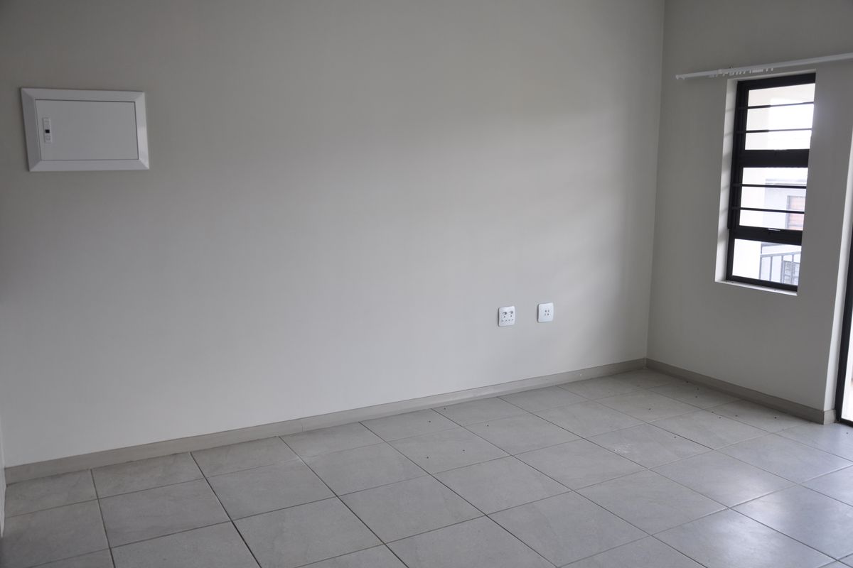 A bright, empty bedroom featuring neutral tiled flooring, a window with security bars, and a wall-mounted electrical distribution board.
