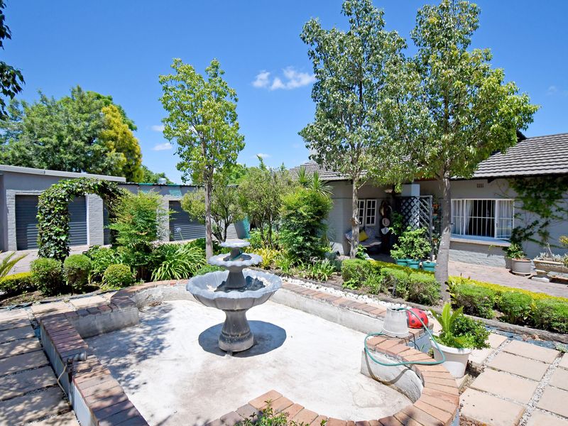 A rear garden area featuring a central stone fountain, brick-edged concrete paving, established trees, and access to the main house and garage structure.