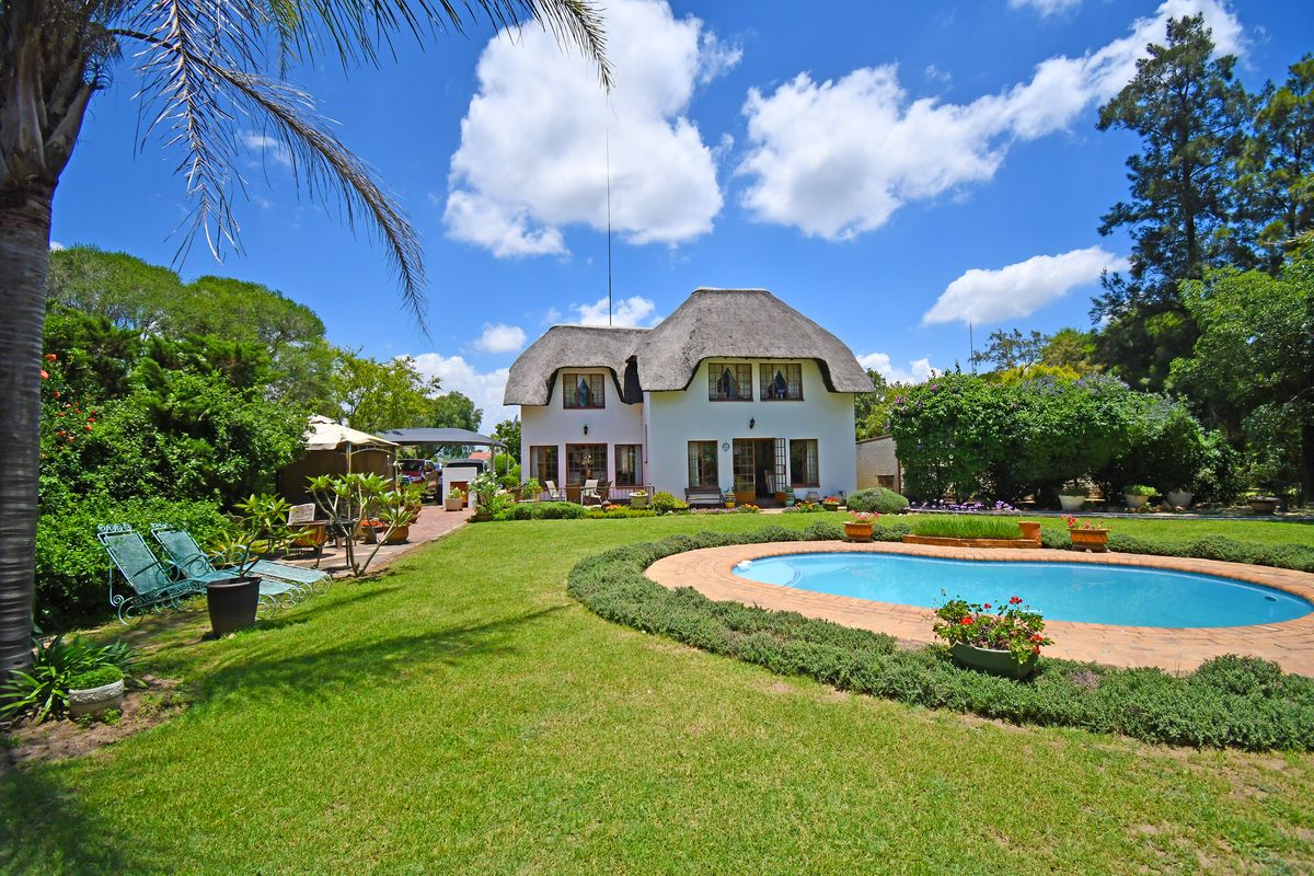Rear view of a charming thatched-roof house featuring a well-maintained garden, a swimming pool with brick coping, and a paved patio area.