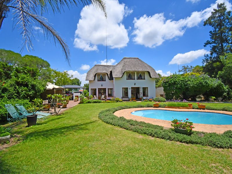 Rear view of a charming thatched-roof house featuring a well-maintained garden, a swimming pool with brick coping, and a paved patio area.