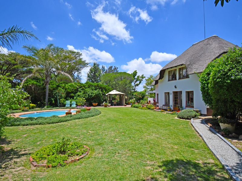A spacious rear garden featuring a well-maintained lawn, a swimming pool, and a charming house with a traditional thatched roof.