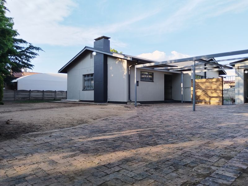 Front exterior view of the house featuring a paved driveway, metal carport structure, and a chimney stack with a surrounding unlandscaped yard.