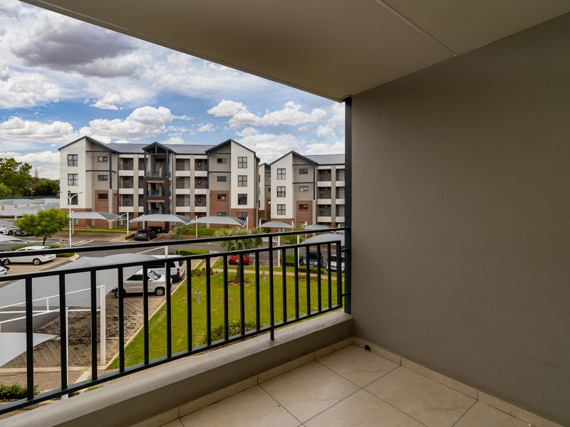A clean, covered balcony featuring tiled flooring and a metal balustrade, offering an elevated view of the apartment complex grounds and parking area.