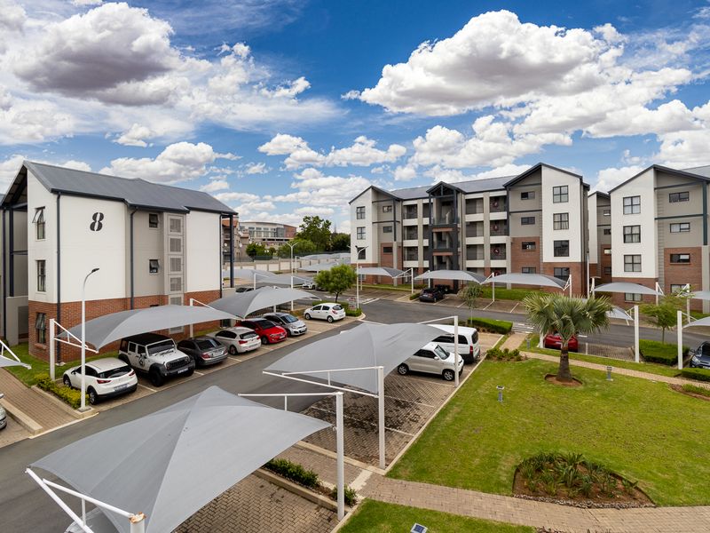 Aerial view of a modern apartment complex featuring multi-story residential buildings, paved parking areas with shade cloth carports, and well-maintained landscaped gardens.