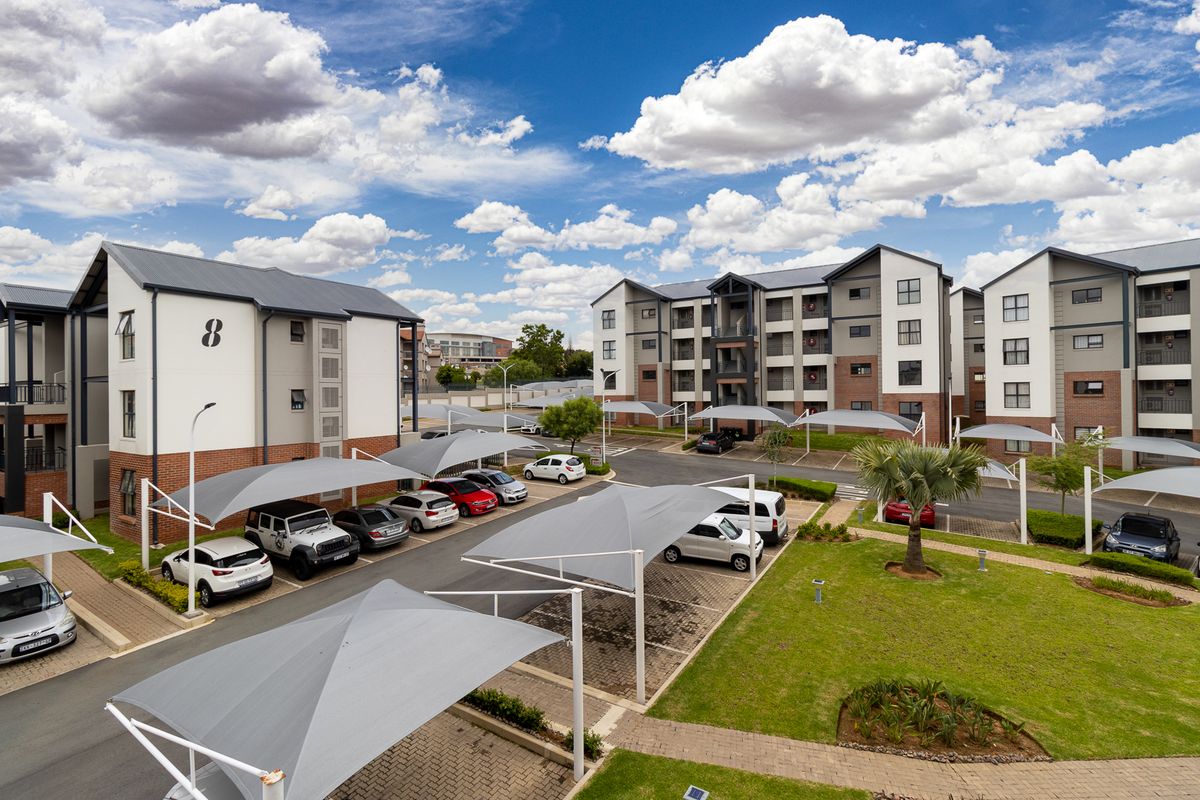 Aerial view of a modern apartment complex featuring multi-story residential buildings, paved parking areas with shade cloth carports, and well-maintained landscaped gardens.