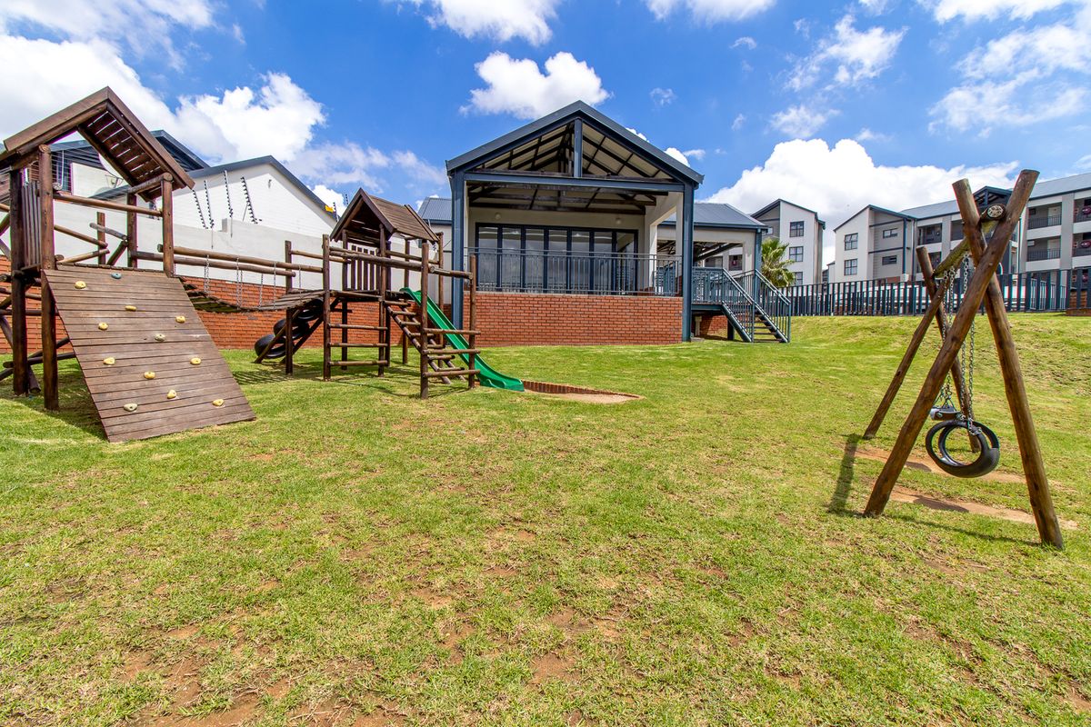 A well-maintained communal garden area featuring a children's playground, manicured lawn, and a covered patio attached to the apartment building.