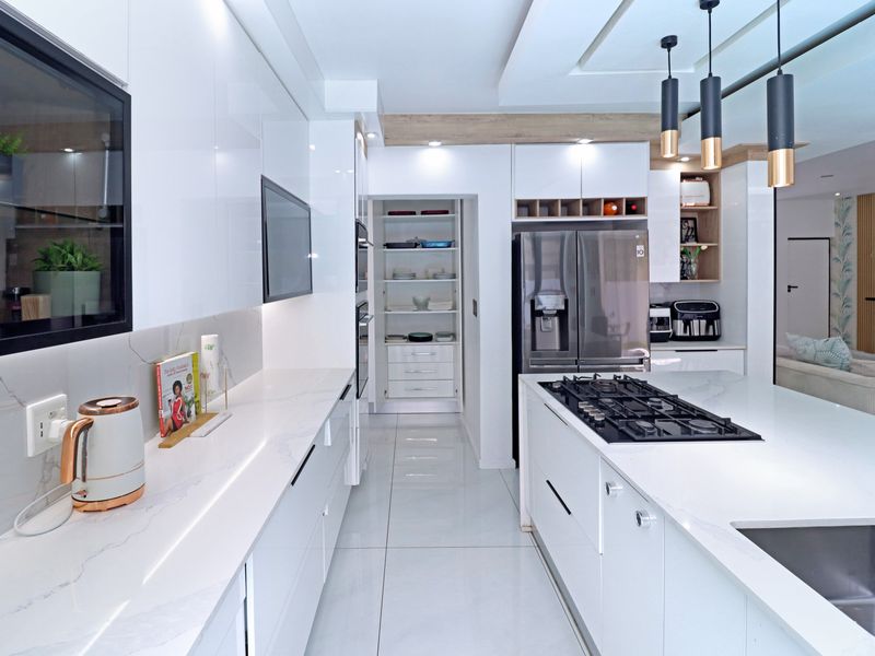 Modern, high-end kitchen featuring sleek white cabinetry, stone countertops, a central island with a gas hob, and an integrated pantry area.