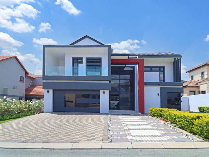 A modern double-story residential property featuring a striking red accent, glass balcony, double garage, and a neatly paved driveway.