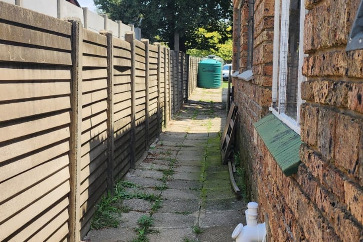 A narrow side exterior walkway featuring brick walling, concrete paving, and a boundary fence, showing signs of wear and maintenance requirements.