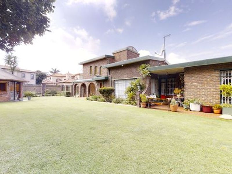 Spacious rear garden featuring a large lawn, brick-faced house with a covered patio area, and a separate thatched outbuilding.