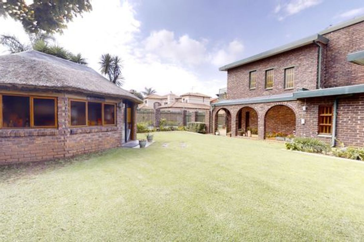 A spacious rear garden featuring a well-maintained lawn, a brick-built main house with an arched patio, and a separate thatched-roof outbuilding.