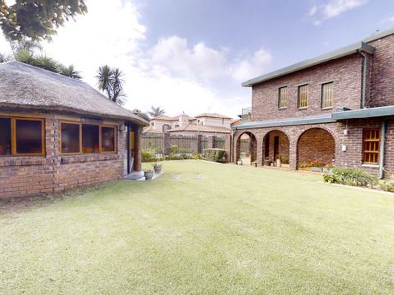 A spacious rear garden featuring a well-maintained lawn, a brick-built main house with an arched patio, and a separate thatched-roof outbuilding.