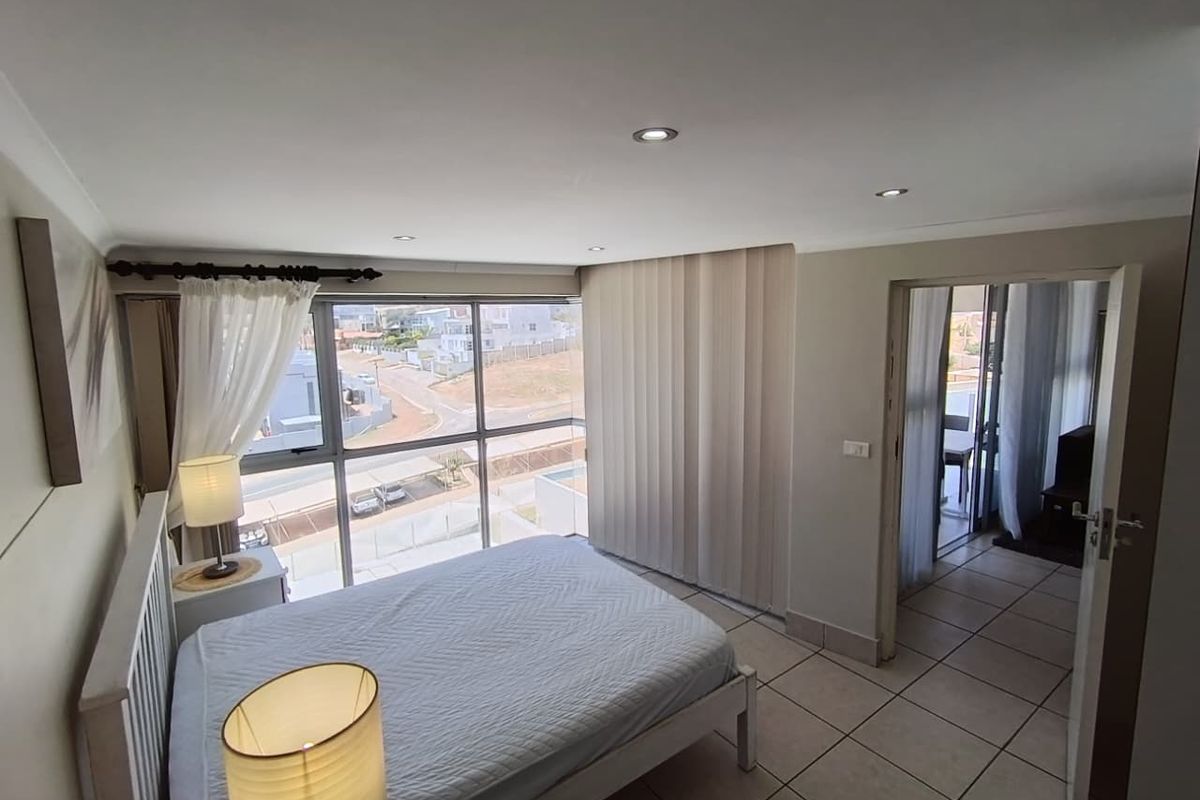 A bright bedroom featuring large floor-to-ceiling windows with neighbourhood views, tiled flooring, recessed ceiling lighting, and direct access to an adjoining balcony area.