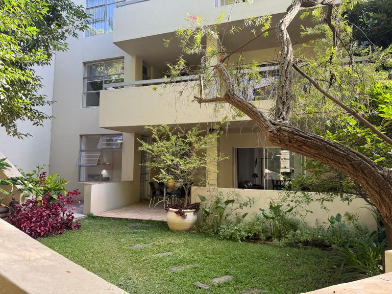 Rear view of the apartment building featuring a ground-floor tiled patio, landscaped garden area, and access to the interior living spaces via sliding doors.