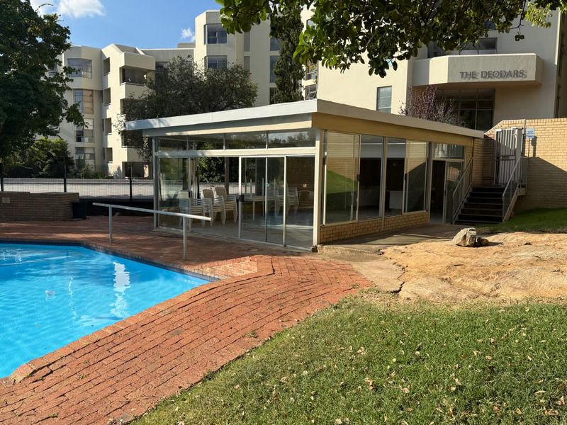 Communal pool area featuring a glass-enclosed clubhouse, brick paving, and landscaped surroundings within a residential apartment complex.