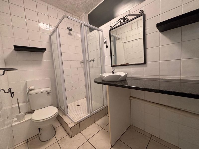 A functional bathroom featuring a shower enclosure, toilet, and a vanity area with a vessel sink, finished with white wall tiles.