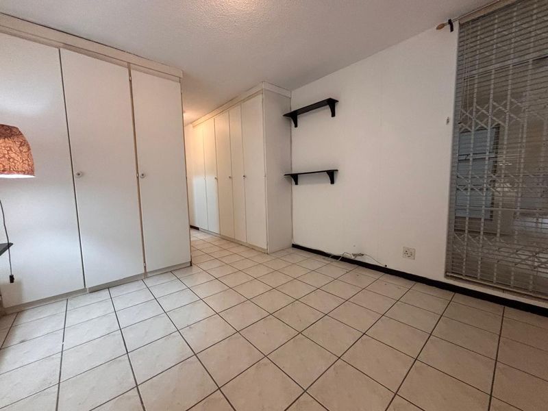 A functional bedroom featuring extensive built-in cupboard storage, neutral tiled flooring, and a secure window with a metal security gate.