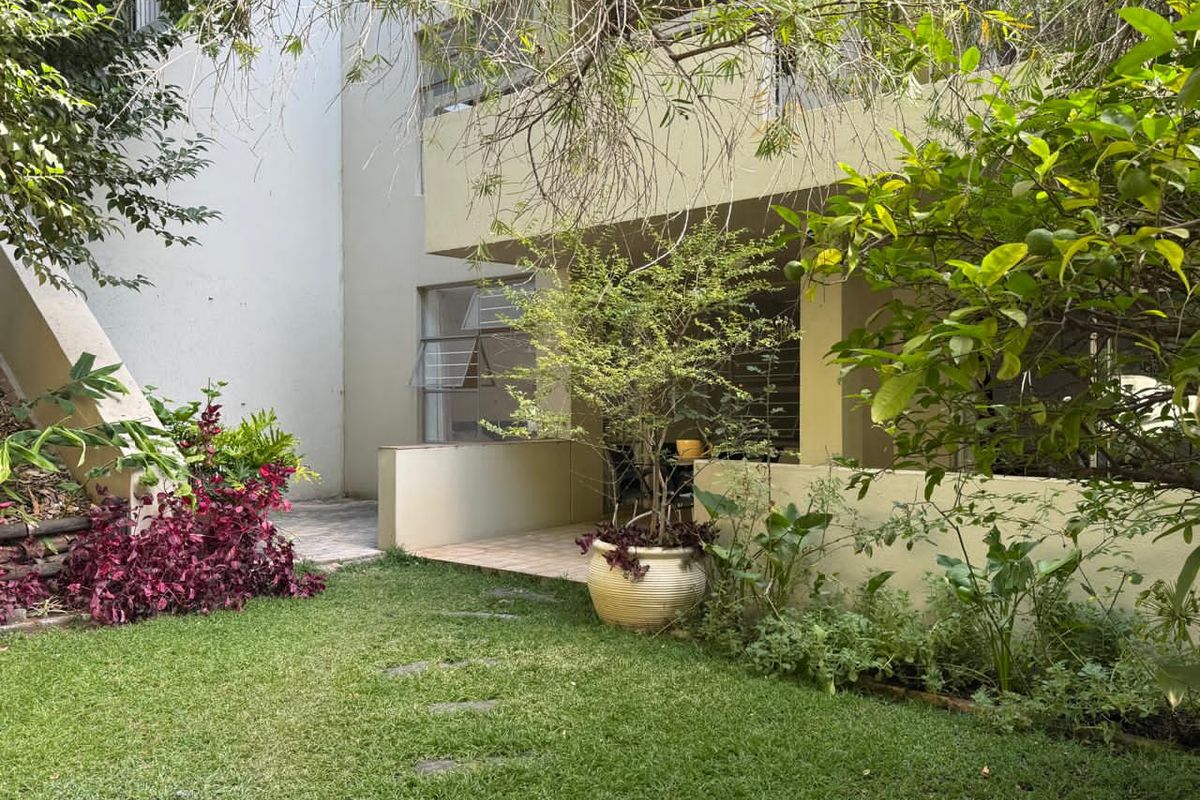 A private rear garden featuring a covered patio area, paved pathway, and established greenery, providing a peaceful outdoor space for the apartment.