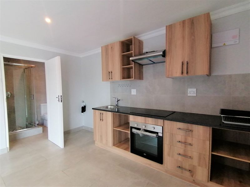 Modern kitchen featuring light wood cabinetry, dark countertops, built-in oven and hob, with an adjacent bathroom visible through the open doorway.
