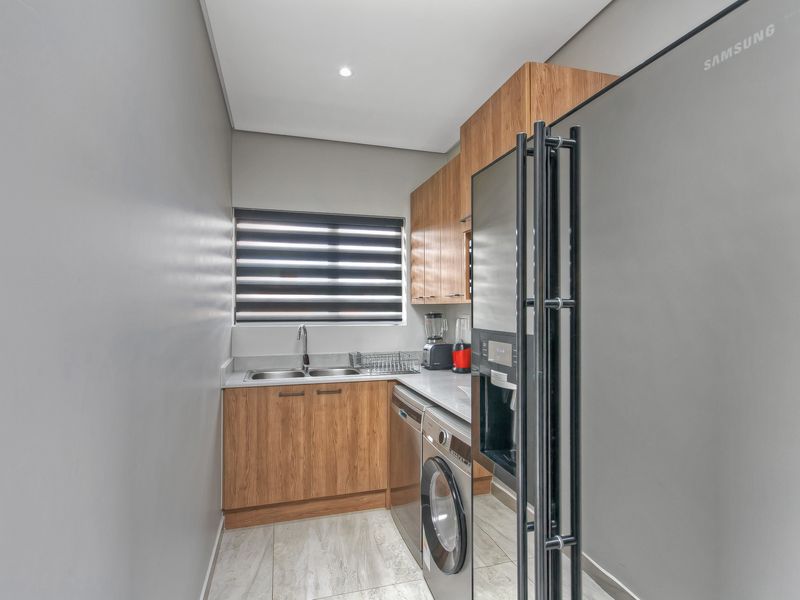 A modern, well-maintained scullery featuring wood-finish cabinetry, stone countertops, a double sink, and tiled flooring, providing efficient additional prep and utility space.