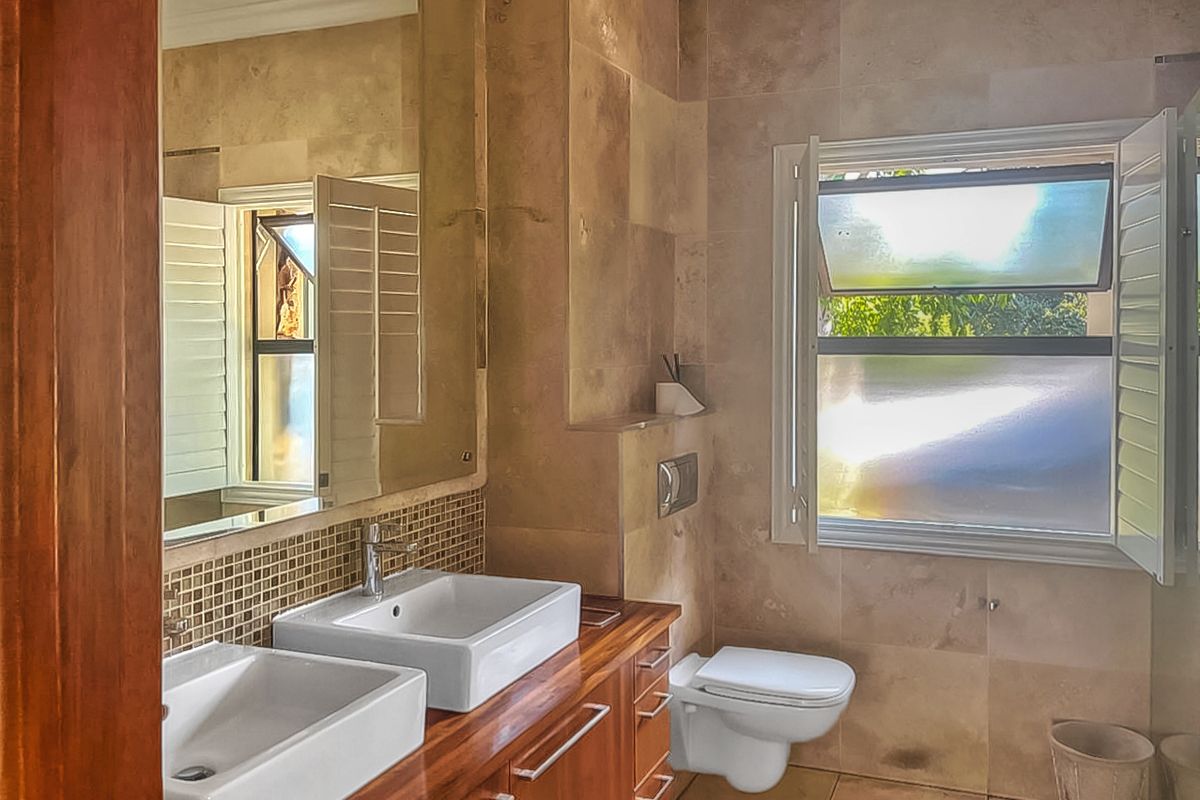 A modern bathroom featuring a double vanity with vessel sinks, wooden cabinetry, wall-mounted toilet, and large window with plantation shutters.