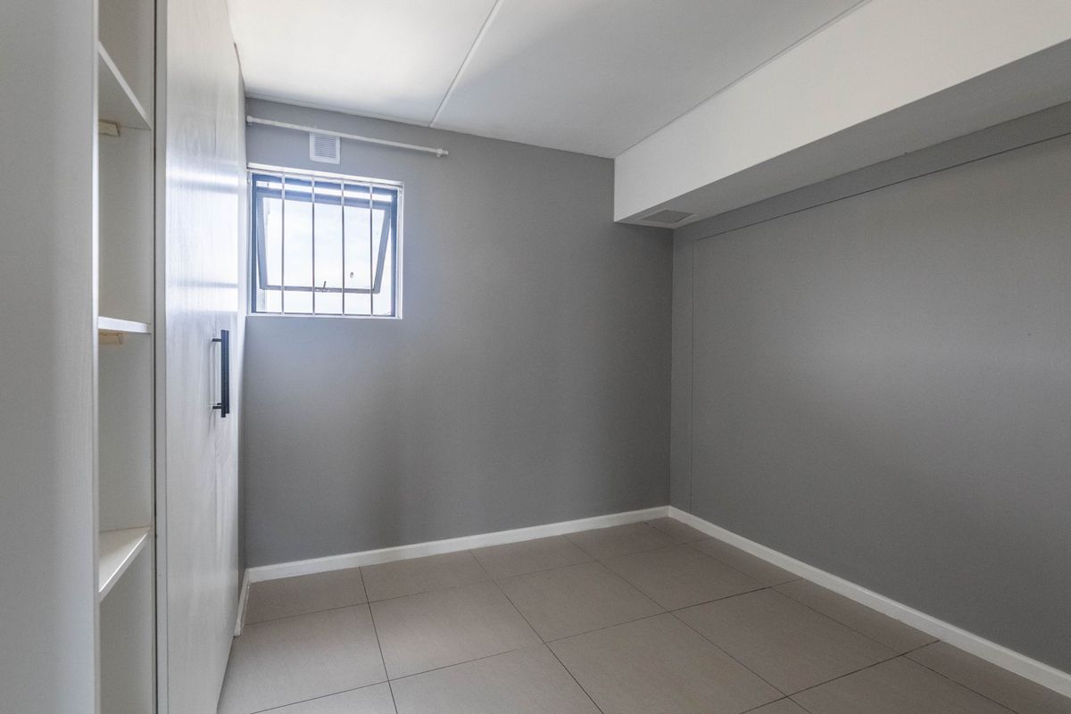 A clean, empty bedroom featuring neutral grey walls, tiled flooring, and built-in storage cabinetry with a small security-barred window.