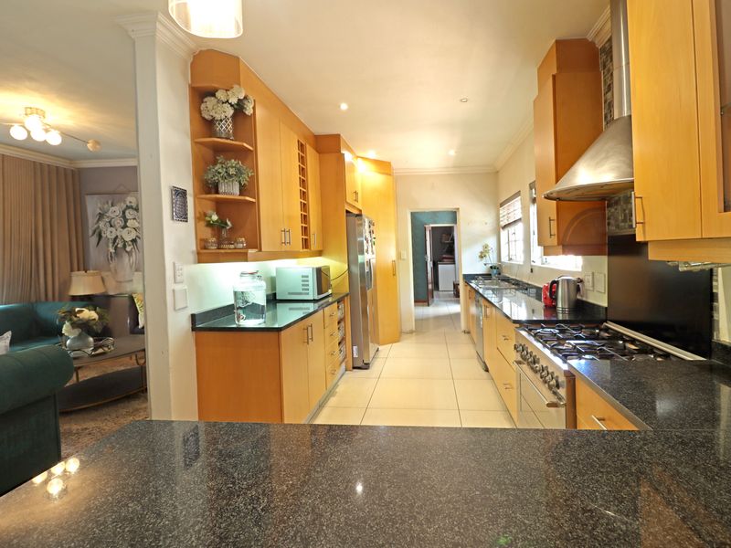 A functional kitchen featuring wooden cabinetry, granite countertops, a gas hob with stainless steel extractor, and tiled flooring, with an adjacent living area.