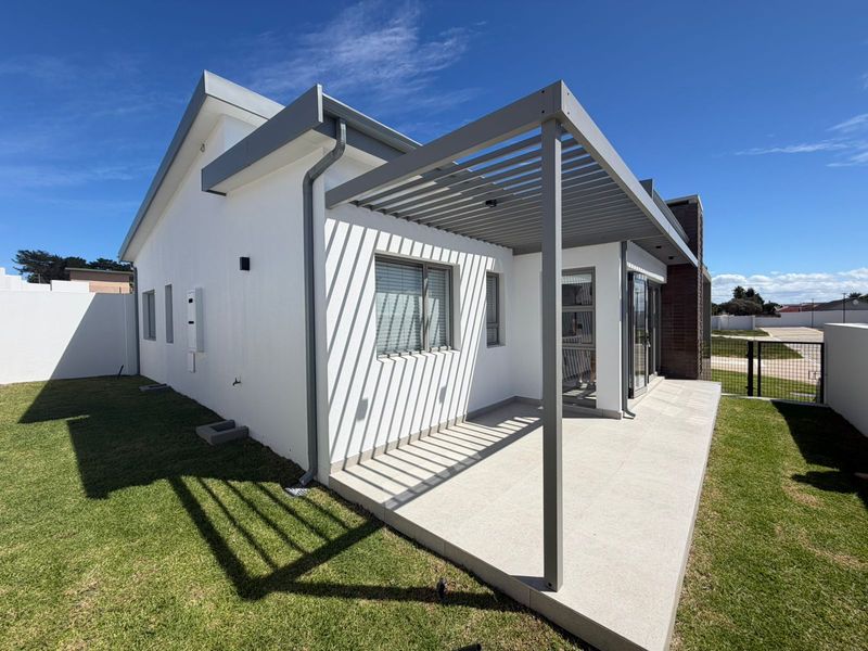 A modern rear exterior view featuring a paved patio area with a contemporary pergola, white rendered walls, and a well-maintained lawn.