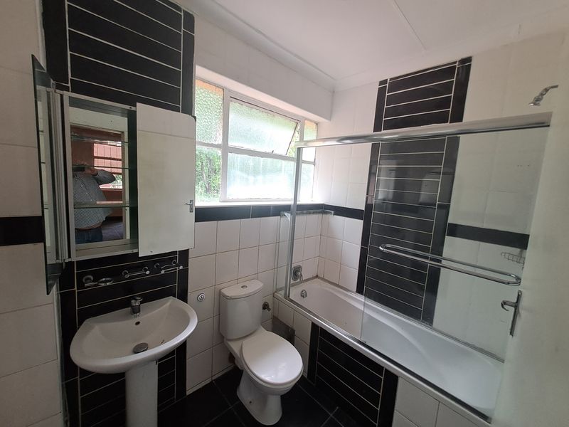 A functional bathroom featuring a pedestal basin, toilet, and a bathtub with a glass shower screen, finished with black and white wall tiling.