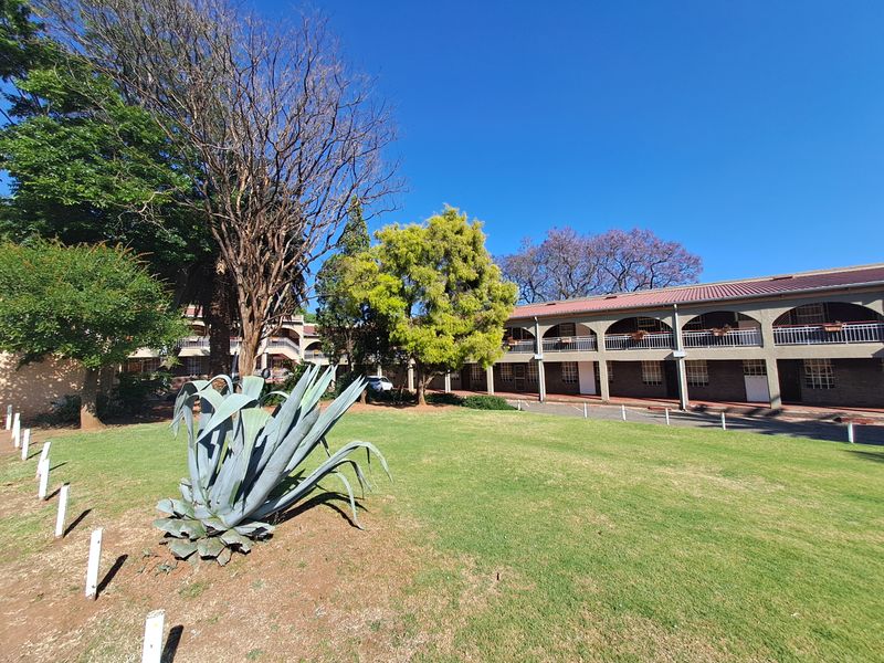 Exterior view of the apartment complex featuring a landscaped garden area, mature trees, and a multi-level building with arched balcony walkways.
