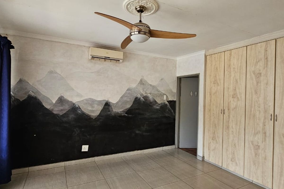 Spacious bedroom featuring built-in light wood cupboards, tiled flooring, a ceiling fan, and an air conditioning unit with a decorative mountain mural wall.