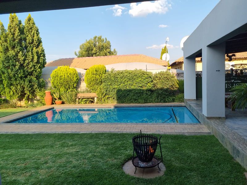 A well-maintained outdoor pool area featuring a clear swimming pool with brick coping, adjacent to a covered patio with concrete pillars and landscaped garden.