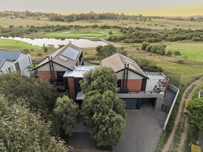 Aerial view of a modern multi-level brick home featuring solar panels, a paved driveway, and a balcony overlooking a golf course and lake.