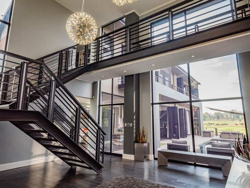 A grand double-volume entrance hall featuring a striking steel staircase, mezzanine walkway, and expansive floor-to-ceiling windows overlooking the pool area.