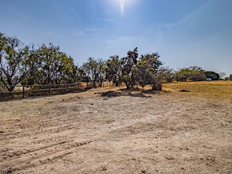 A view of a vacant residential plot featuring dry grass, mature trees, and a wooden post and rail boundary fence.