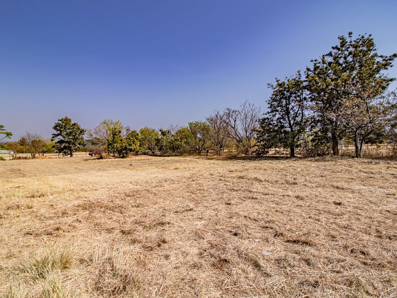 A wide-angle view of a vacant residential plot featuring dry grass, mature trees, and perimeter fencing under a clear blue sky.