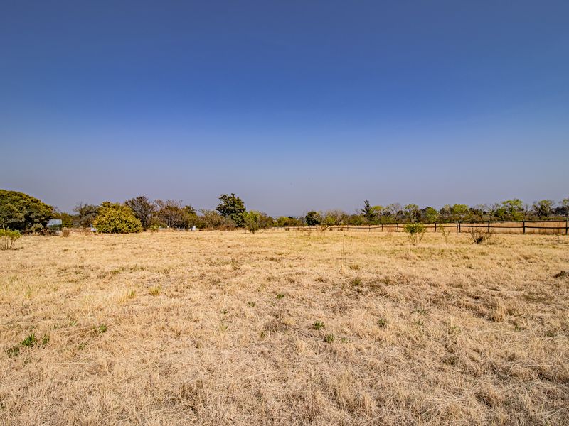 A wide-angle view of a vacant residential plot featuring dry grass, mature trees, and perimeter post and rail fencing.