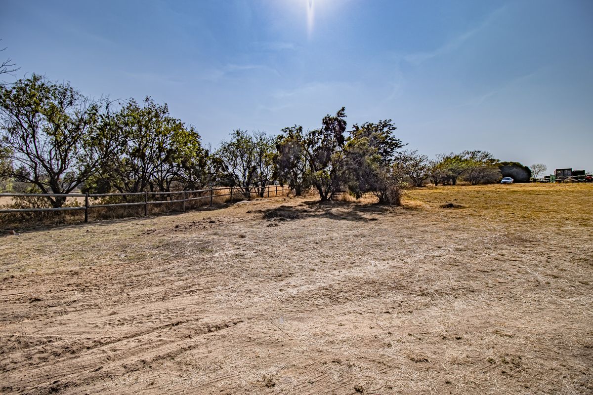 A view of a vacant residential plot featuring dry grass, mature trees, and a wooden post and rail boundary fence.