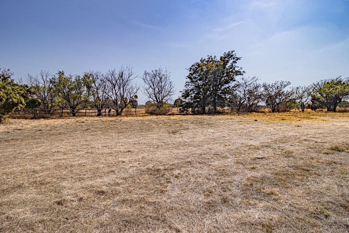 A wide-angle view of a vacant residential plot featuring dry grass, mature trees, and a perimeter wire fence.