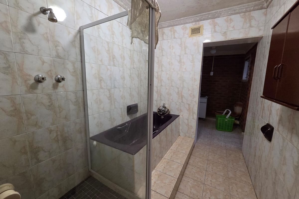 A dated bathroom featuring a built-in bathtub, glass shower screen, and full-height wall tiling. The space requires cosmetic updates to modernize the finishes.