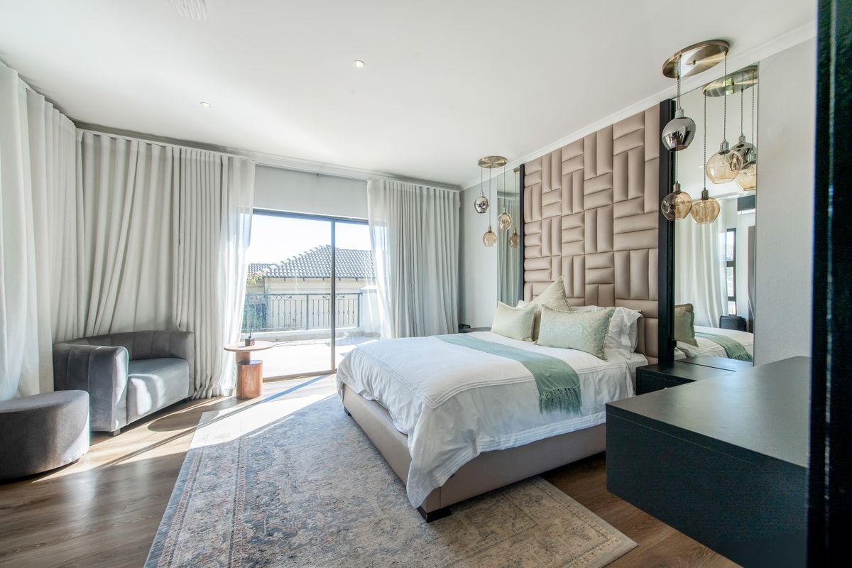 A modern, well-appointed bedroom featuring custom upholstered wall paneling, recessed lighting, and a sliding glass door leading to an outdoor balcony.