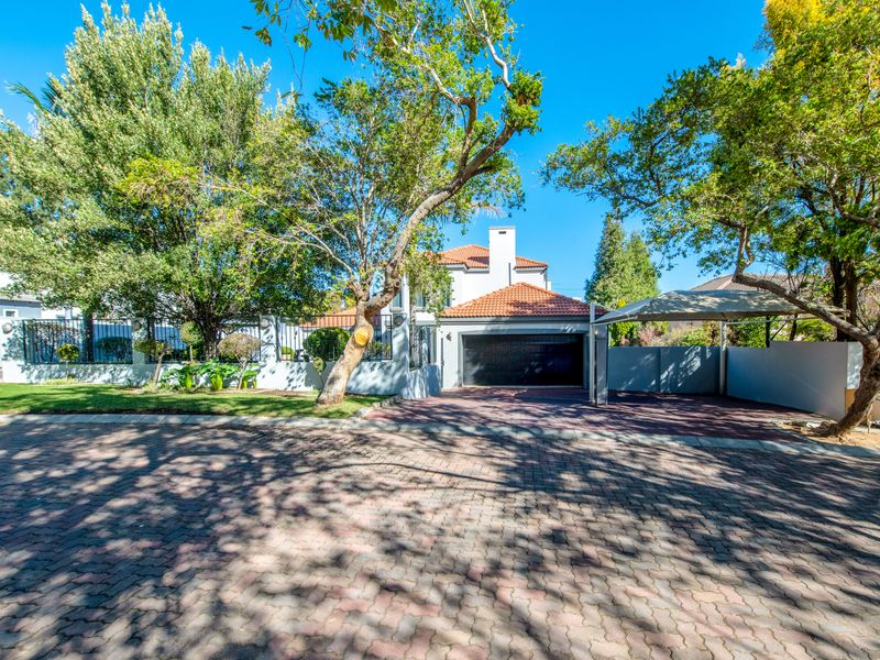 Front view of the property featuring a paved driveway, double garage, additional carport, and established garden with mature trees.