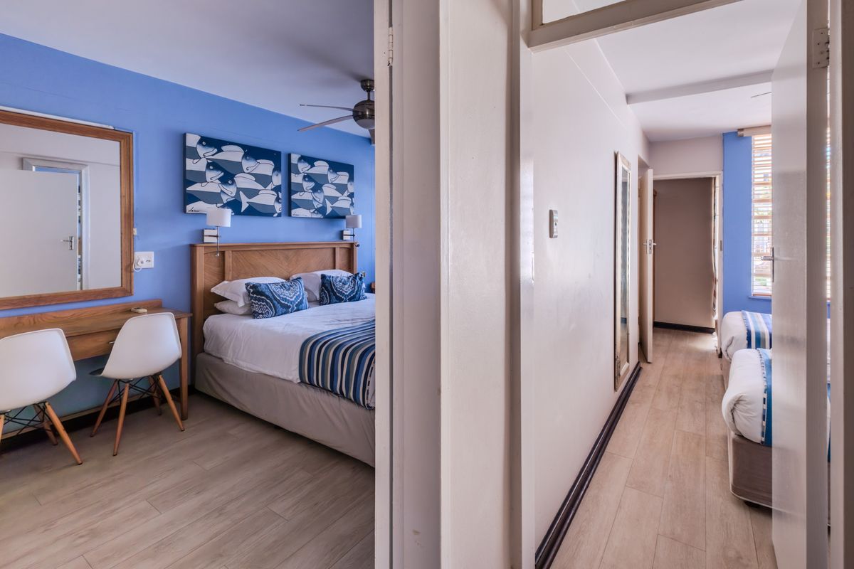 A well-maintained bedroom featuring light laminate flooring, a ceiling fan, and a built-in vanity area, with an adjacent hallway leading to additional sleeping space.