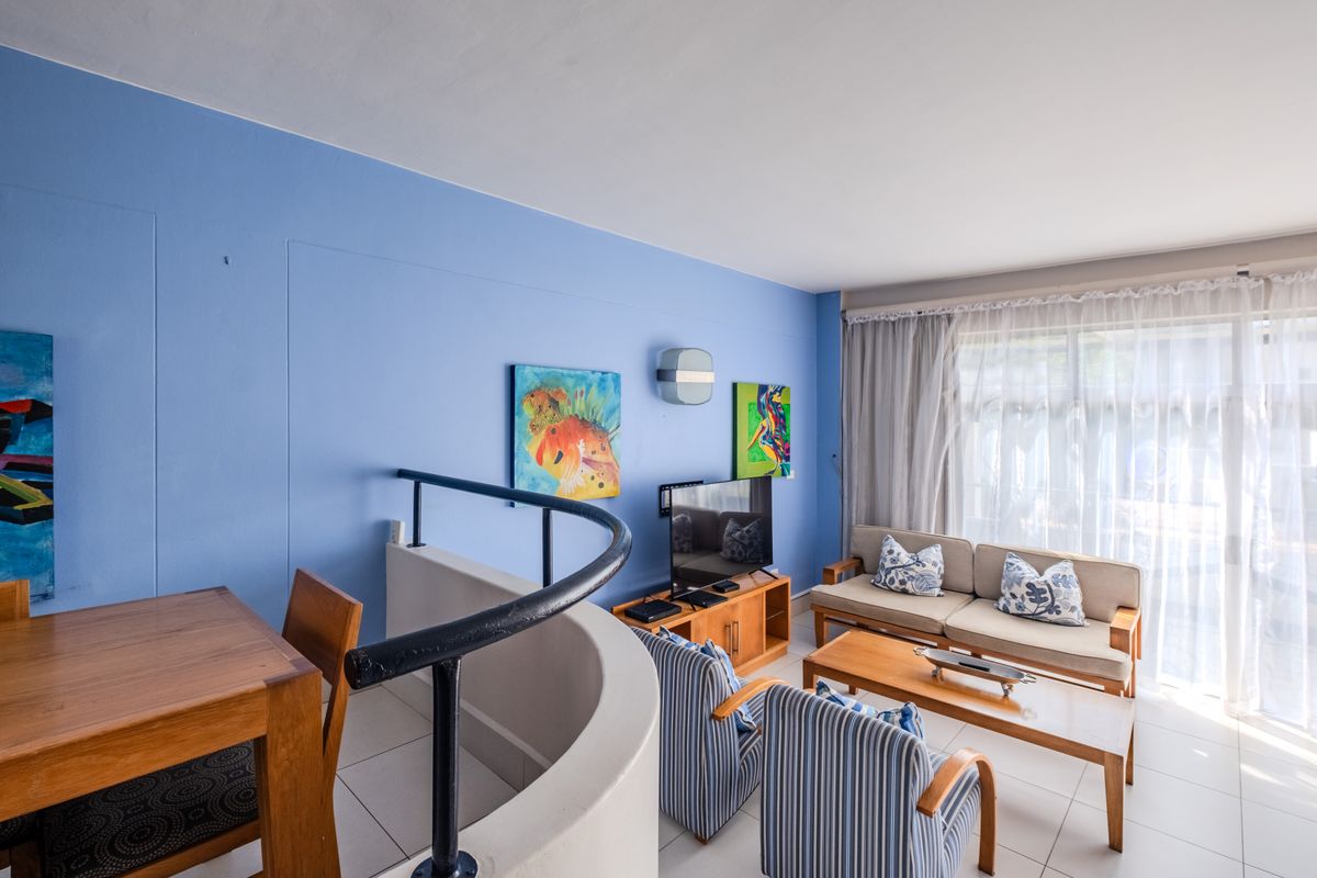 A bright living room featuring tiled flooring, a curved staircase balustrade, and large windows with curtains, offering a functional and open living space.