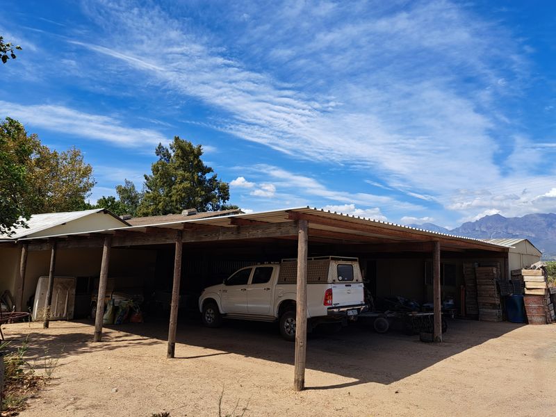 An open-sided farm carport or storage structure featuring a corrugated metal roof supported by timber posts, providing sheltered parking for vehicles and equipment.